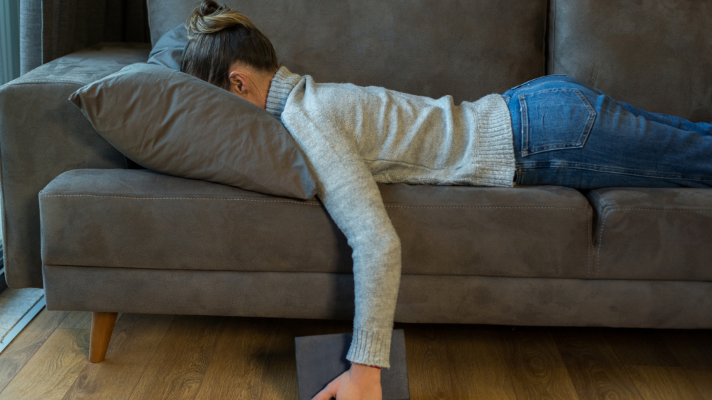 A woman with hair pinned up in bun, wearing a sweater and jeans, lies face down on the sofa.